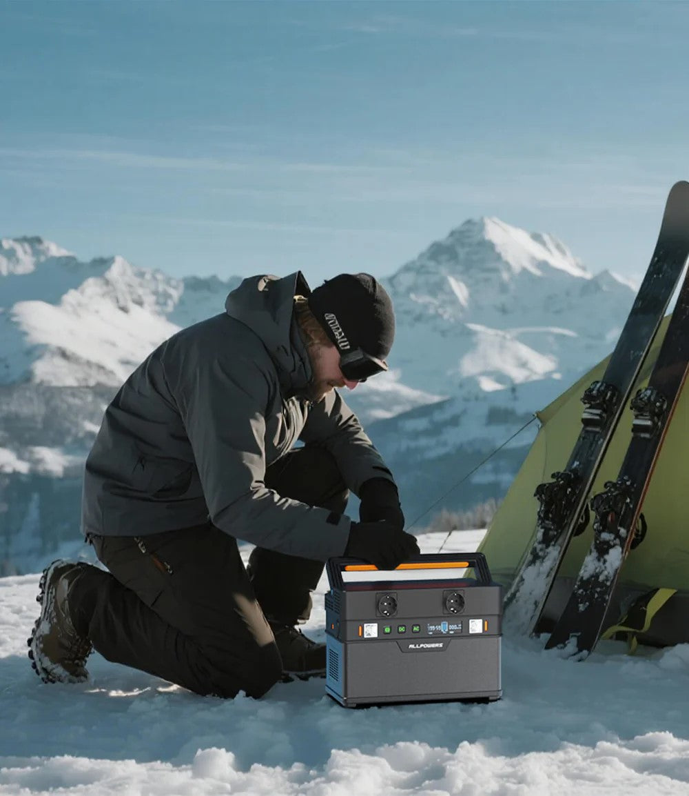Um homem agachado na neve, trabalhando com uma estação de energia portátil em uma cena de aventura outdoor no inverno, com montanhas cobertas de neve ao fundo e esquis ao lado.